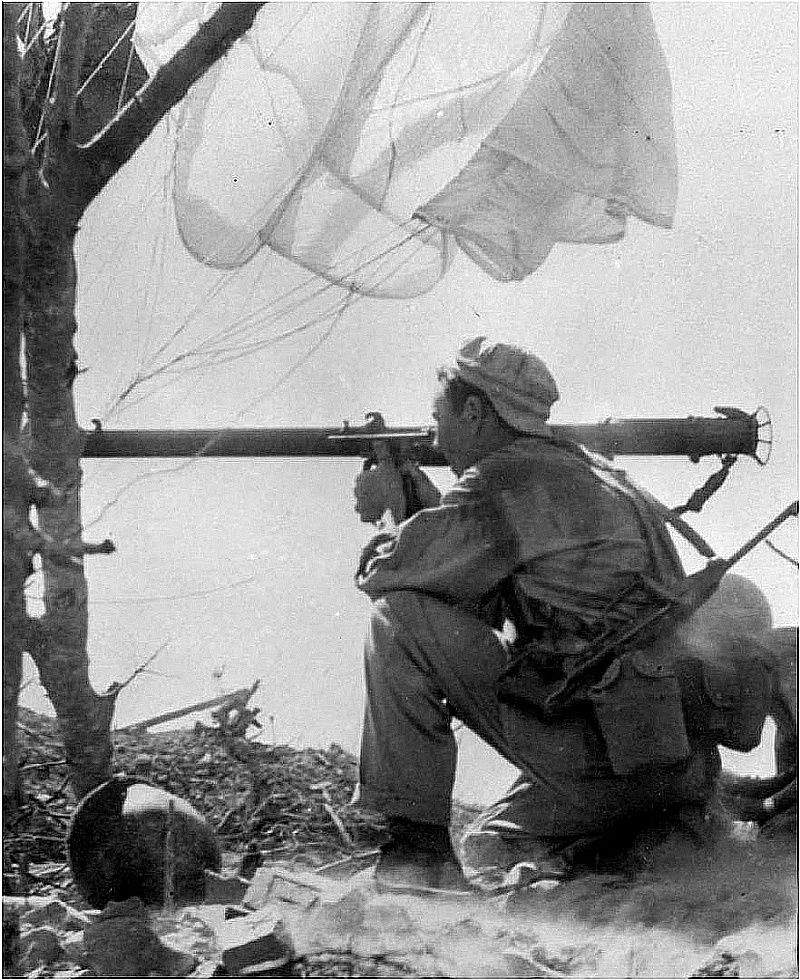 An American paratrooper fires a bazooka with an M-1A1 slung across his back.