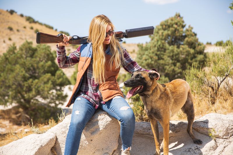 Woman petting a dog with one hand while the other hand balances a Remington 870 with SilencerCo Salvo-12 suppressor on her shoulder.