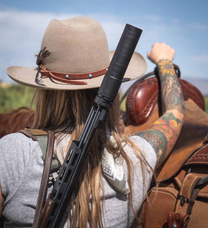 Woman with suppressed rifle slung over shoulder in front of a saddled horse