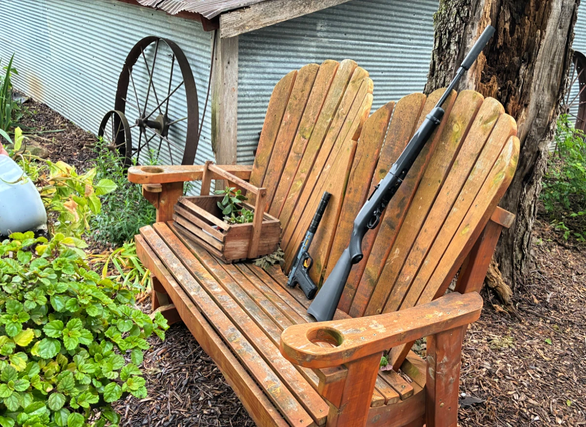 two suppressed rimfire guns on a wooden bench in a country garden